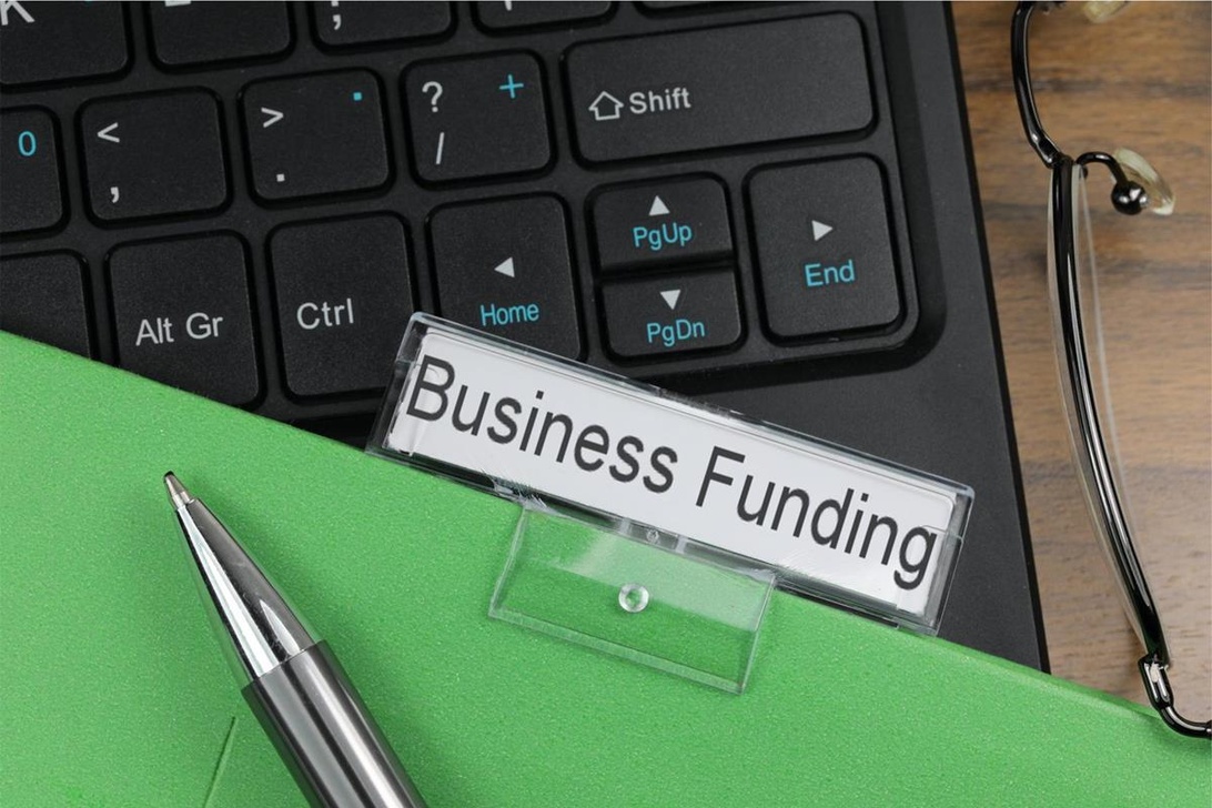 En grön mapp med etiketten 'Business Funding' på ett laptop-tangentbord med penna och glasögon i närheten, vilket symboliserar organisation av information kring företagsfinansiering.