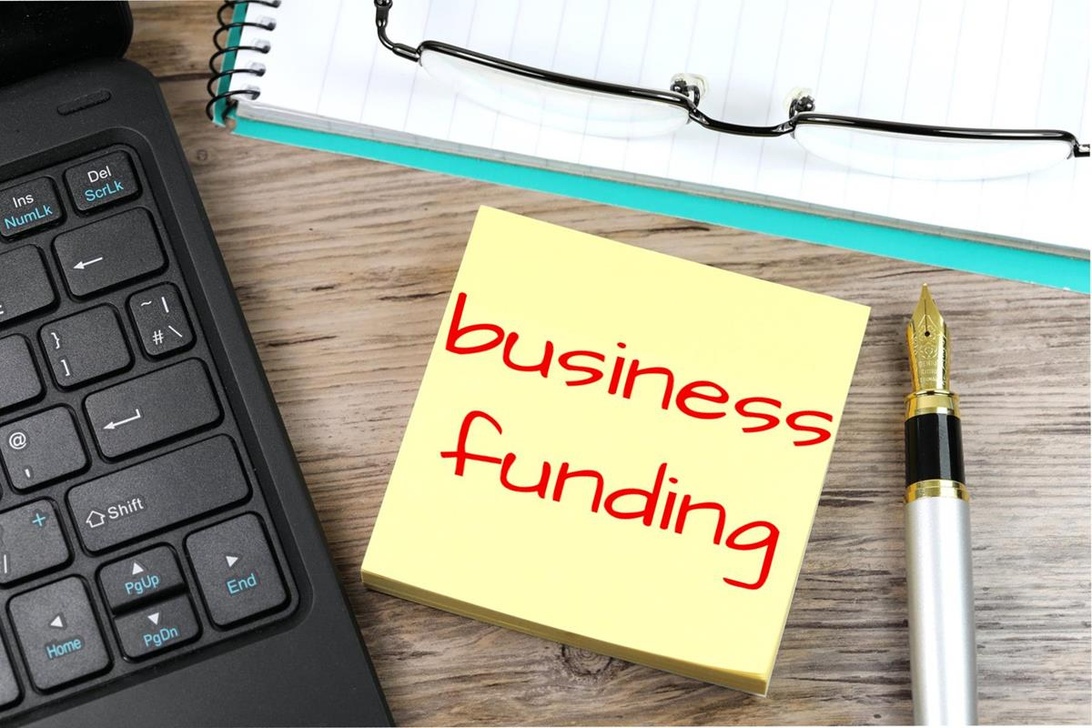 En gul kom-ihåg-lapp med texten 'business funding' handskrivet med röd penna, placerad på ett laptop-tangentbord.