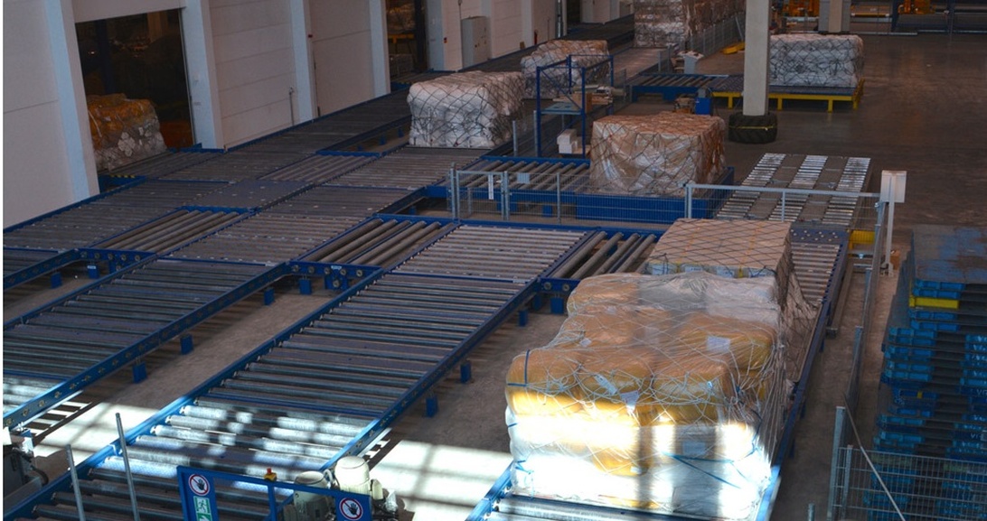 Interiör av en logistikanläggning med rulltransportörer och paketerat gods, som visar materialhanteringsinfrastruktur för lagerdrift.